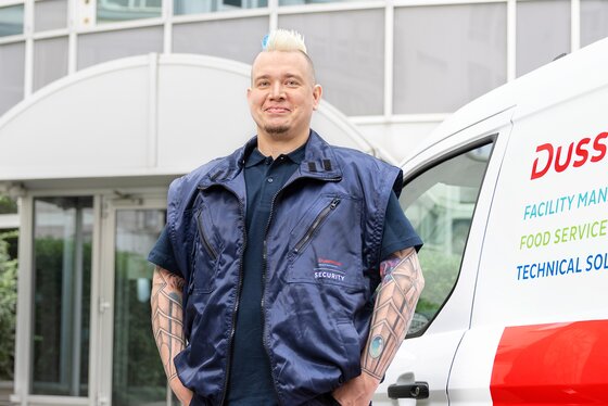 Dussmann - Profi des Alltags Maik RIchter Mitarbeiter vom Sicherheitsdienst Dussmann mit ARbeitskleidung vor einem Dussmann-Fahrzeug stehend